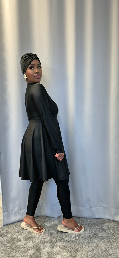 A woman models the Shop Stylish Black Burkini by Bihi Boutique, featuring a long-sleeve dress, matching leggings, and a black headwrap, standing sideways before a gray curtain and looking over her shoulder at the camera.
