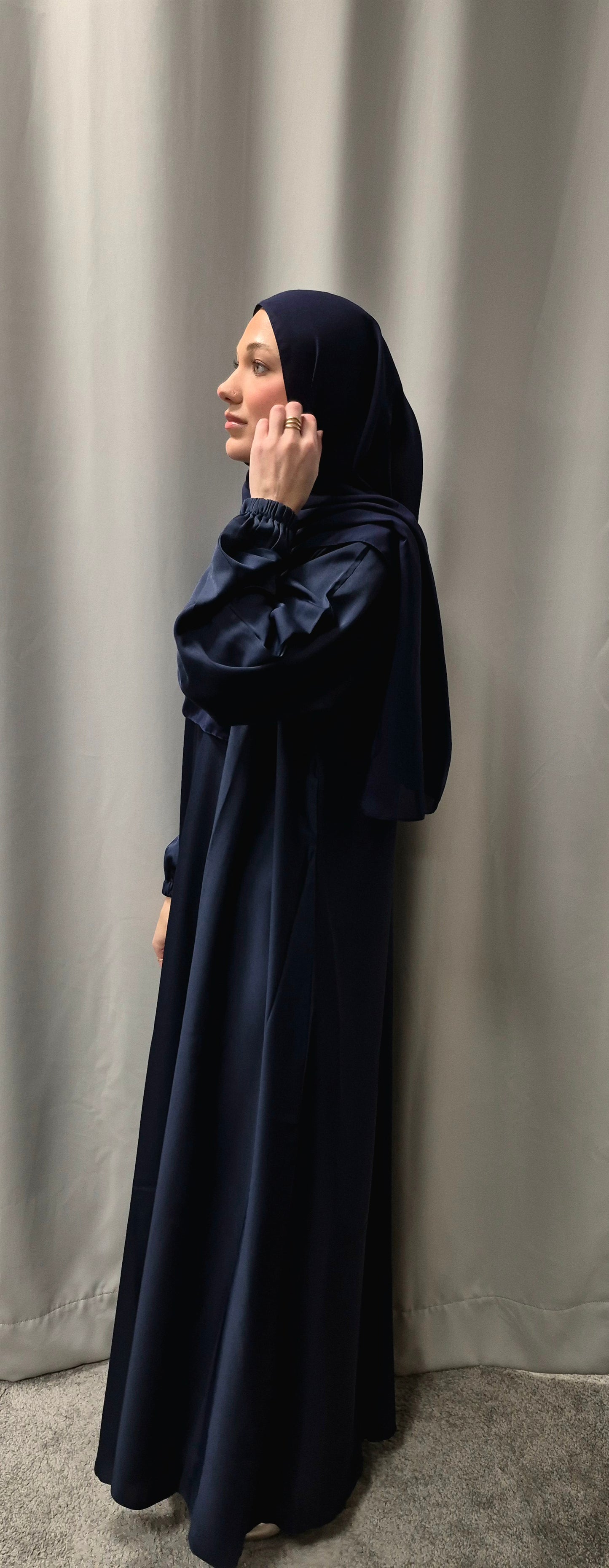 A woman models the Stylish Navy Blue Abaya with Chiffon Hijab (Size 58) by Bihi Boutique, standing in profile against a light gray curtain with her hand near her face.
