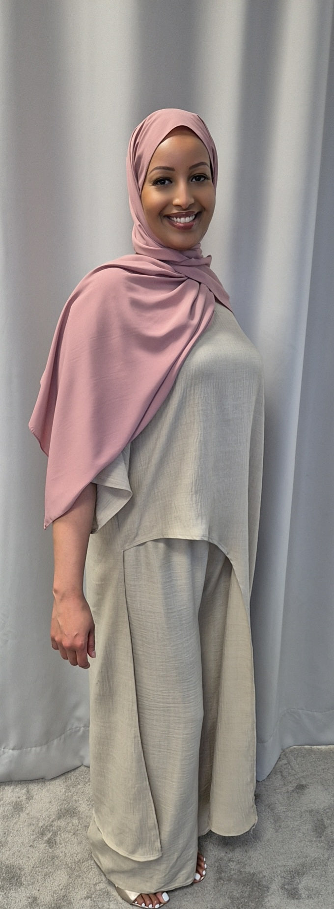 A woman smiles in front of gray curtains, wearing the Shop Luxurious 2-Piece Linen Set in light beige and a pink hijab from Bihi Boutique.