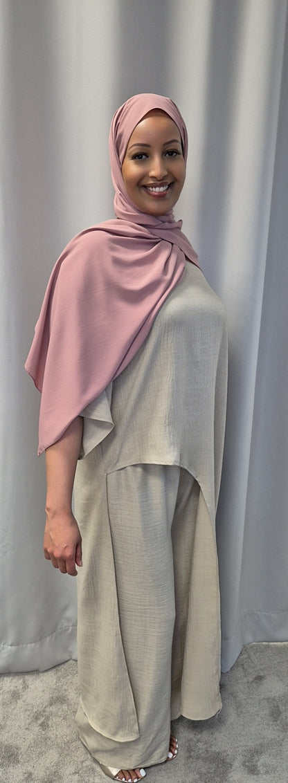 A woman smiles in front of gray curtains, wearing the Shop Luxurious 2-Piece Linen Set in light beige and a pink hijab from Bihi Boutique.