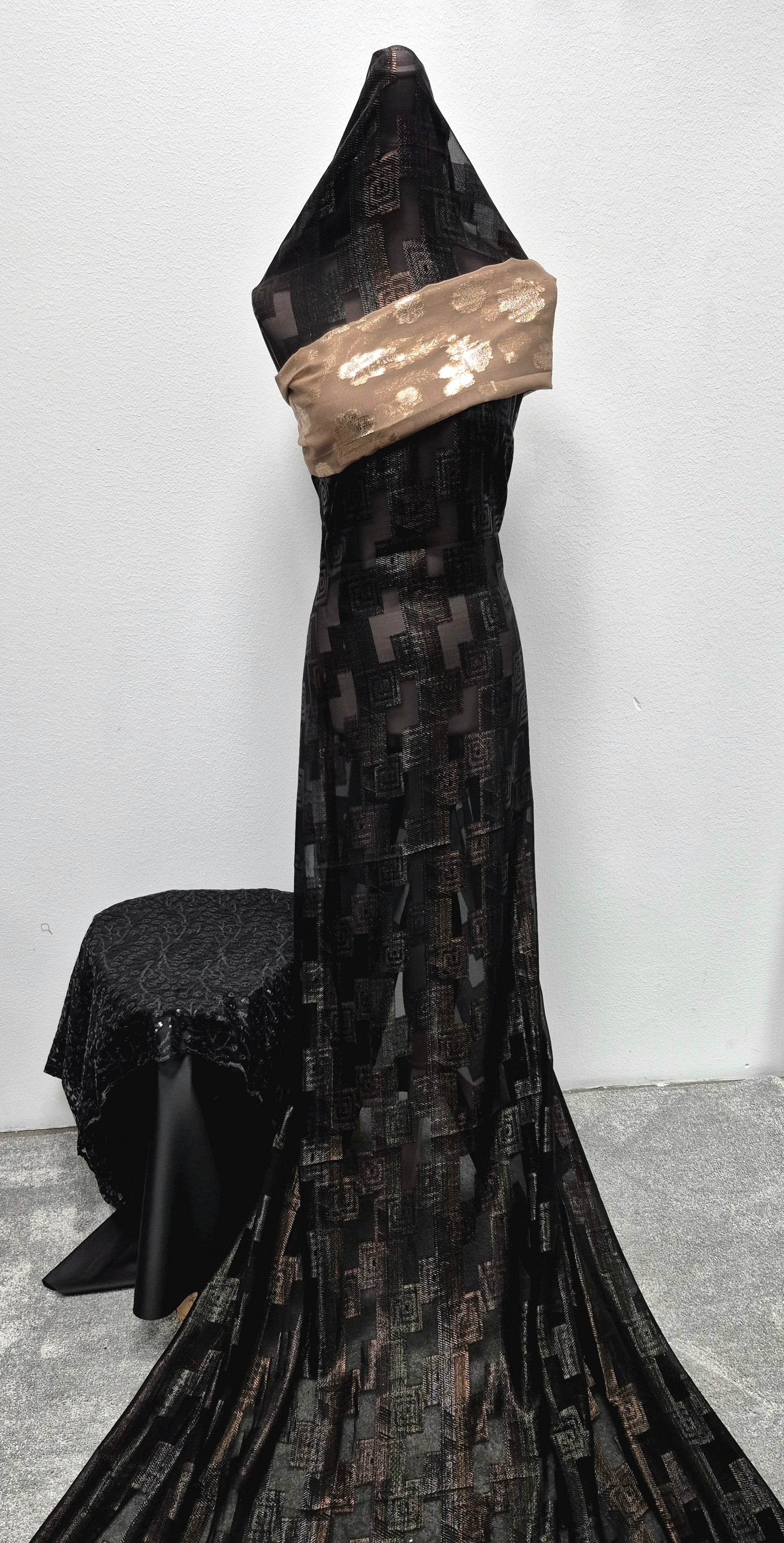 A mannequin displays the Shop Stylish Fransawi Dirac Bacweyne dress by Bihi Boutique—an elegant, flowing black piece with geometric patterns and a shiny beige sash—next to a matching table, all set against a plain white wall.