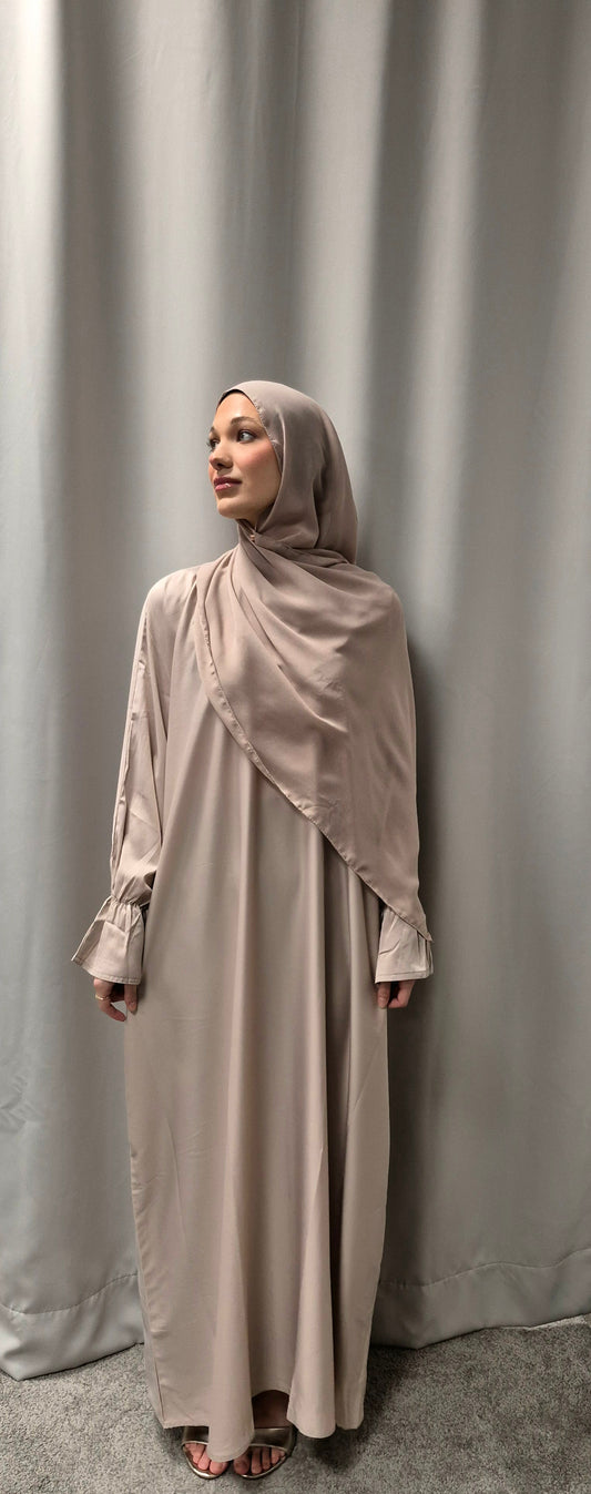 A woman models the Stylish Marwa Abaya Size 54 from Bihi Boutique, featuring elegant and trendy designs in light beige, standing barefoot before a light grey curtain and gazing calmly to her left.