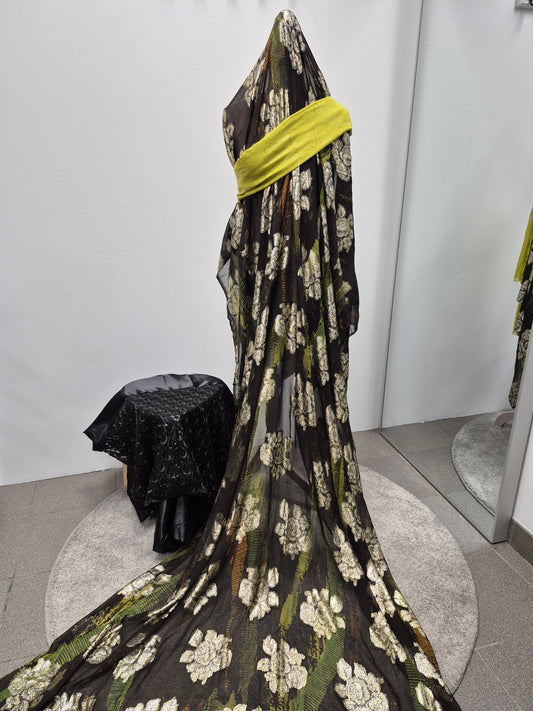 A mannequin displays the Buy Fransawi Dirac Half Bacloth from Bihi Boutique—sheer black fabric with white floral patterns and a yellow sash—standing on a round mat near a wall, mirror, and small covered table.