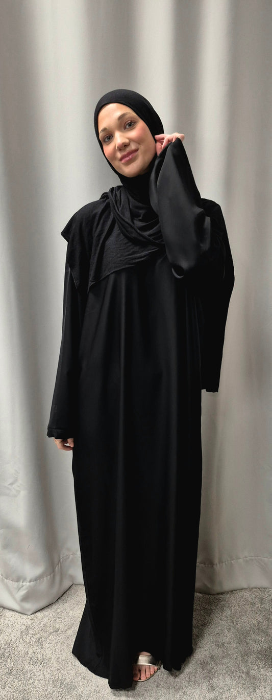 A woman smiles softly at the camera, barefoot on a light carpet in front of a plain curtain, wearing the Shop Layla Closed Abaya - Classic Black Everyday Elegance from Bihi Boutique with a matching hijab.
