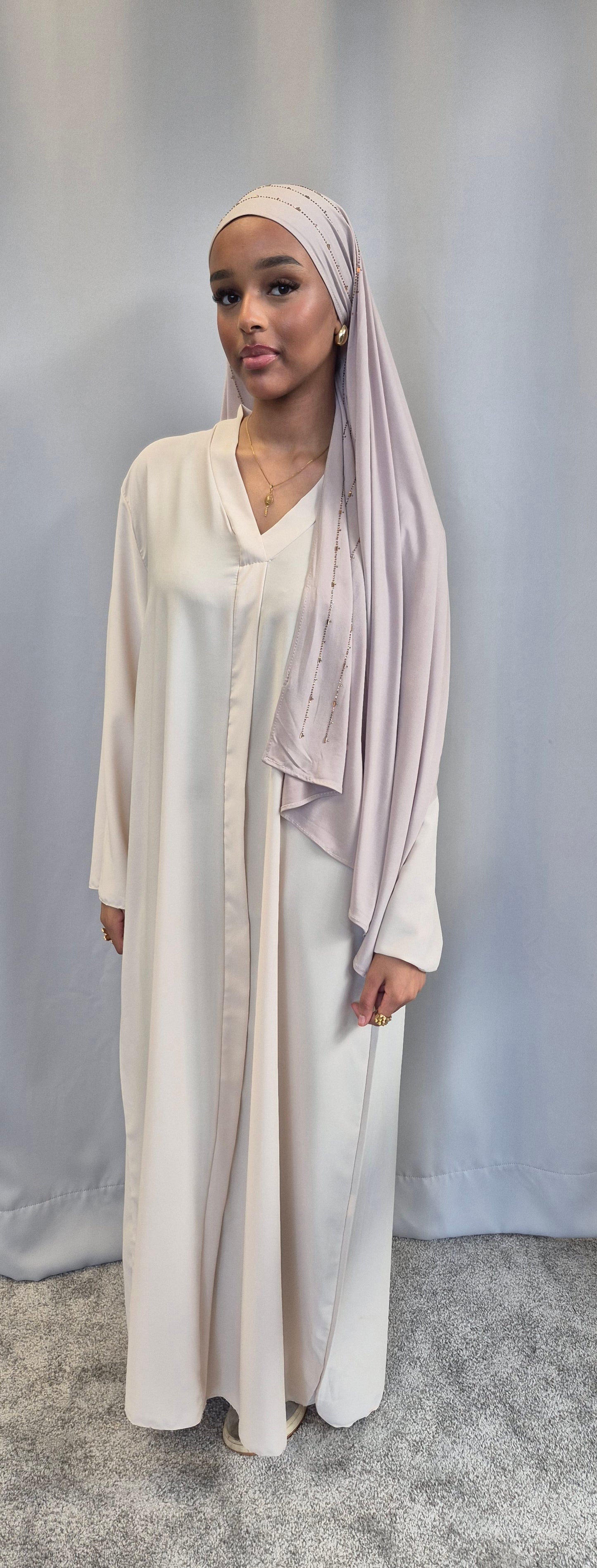 A woman in the Islene Dress by Bihi Boutique (available in 3 colors, fits 160-165 cm) and a light pink hijab stands before a plain backdrop, accessorized with gold jewelry and wearing a calm expression.