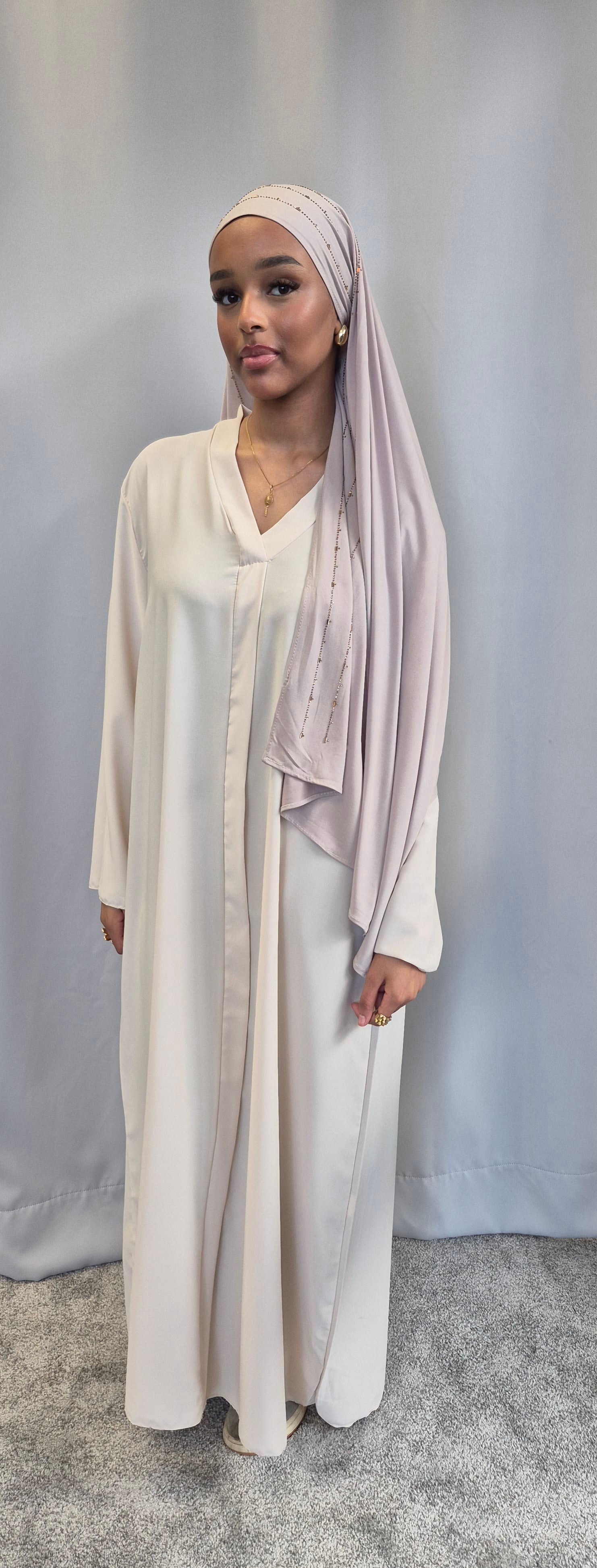 A woman in the Islene Dress by Bihi Boutique (available in 3 colors, fits 160-165 cm) and a light pink hijab stands before a plain backdrop, accessorized with gold jewelry and wearing a calm expression.