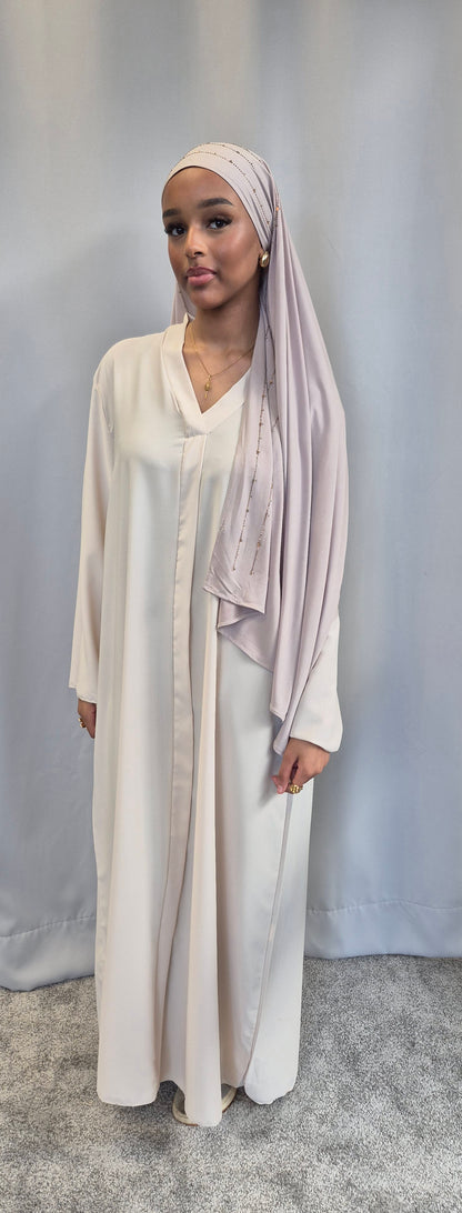A woman in the Islene Dress by Bihi Boutique (available in 3 colors, fits 160-165 cm) and a light pink hijab stands before a plain backdrop, accessorized with gold jewelry and wearing a calm expression.