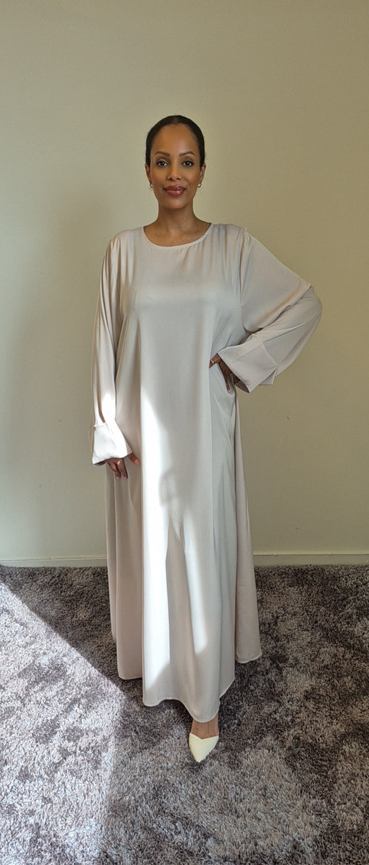 A woman poses indoors on a carpeted floor, wearing the Elegant Satin Abaya Noireen by Bihi Boutique—an evening dress with wide sleeves, pockets, and cream shoes—standing with one hand on her hip against a plain wall.