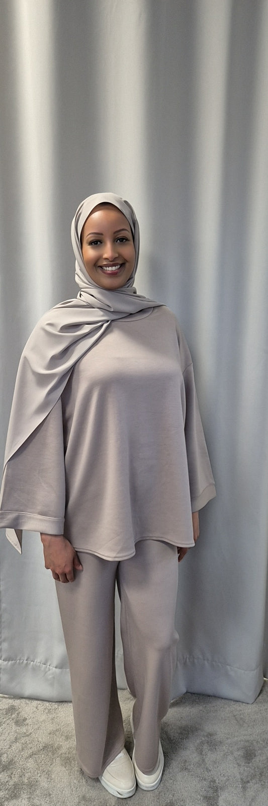 A smiling woman poses in front of a light grey curtain, wearing Bihi Boutique’s Calma Luxe Nue & Noir one-size-fits-all set—a stylish, chic matching hijab, tunic, and pants—paired with white slip-on shoes.
