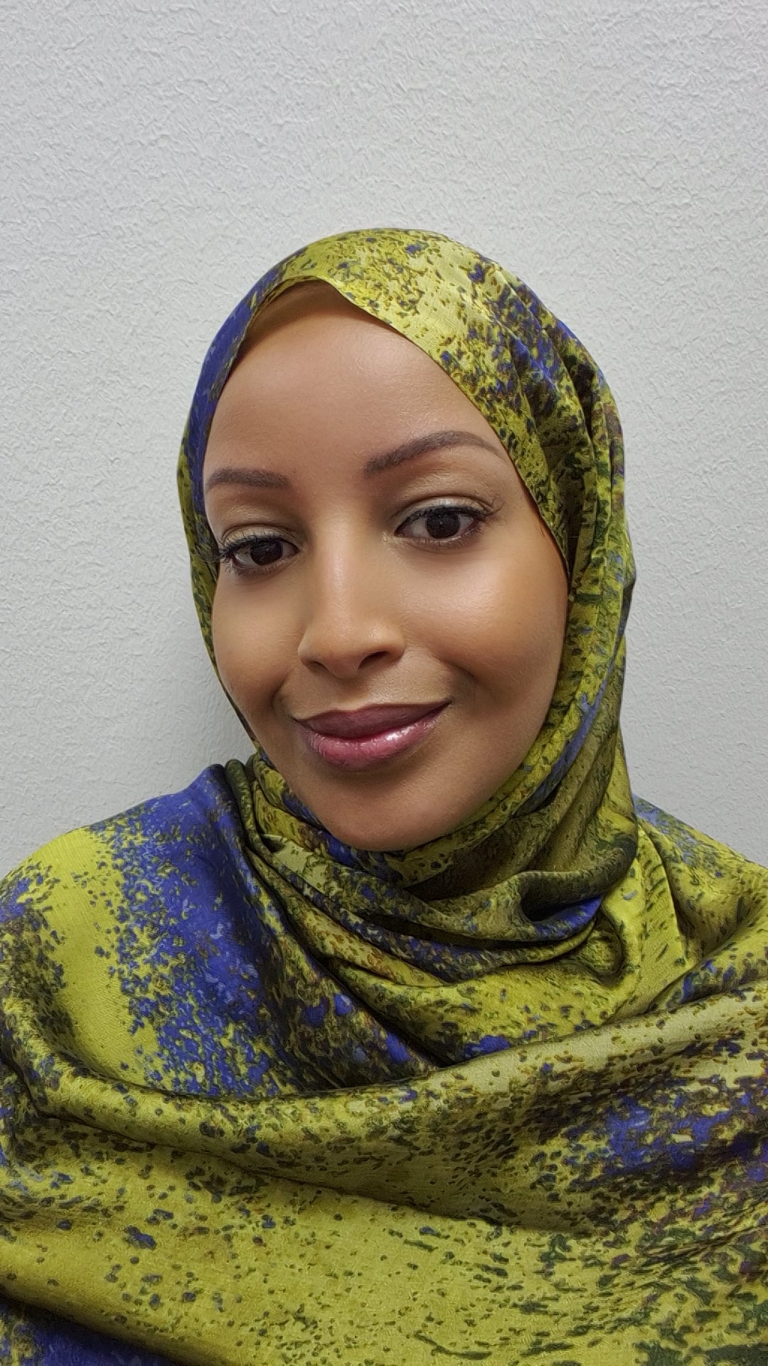 A woman smiles gently at the camera, wearing the Trendy Printed Chiffon Hijab by Bihi Boutique—a stylish and lightweight scarf with a yellow and blue pattern—set against a light textured background.