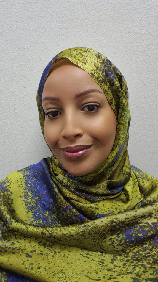 A woman smiles gently at the camera, wearing the Trendy Printed Chiffon Hijab by Bihi Boutique—a stylish and lightweight scarf with a yellow and blue pattern—set against a light textured background.