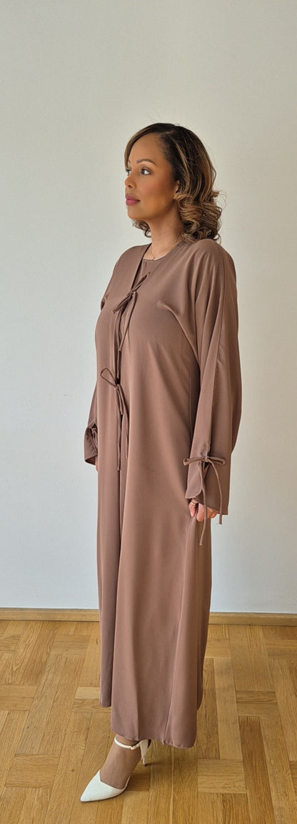 A woman stands indoors on a wooden floor, wearing the Bihi Boutique Stylish Ayan Brown Hijab Set in luxurious Nidah & Chiffon with ties at the sleeves and front, paired with white pointed-toe heels. She faces left, looking slightly upward.