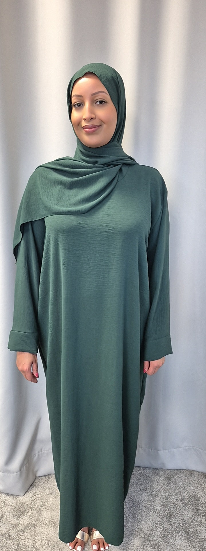 A woman stands in front of a light grey curtain, wearing the Elegant Black One Size Prayer Clothing by Bihi Boutique, suitable for all sizes. She smiles gently with her hands relaxed at her sides.
