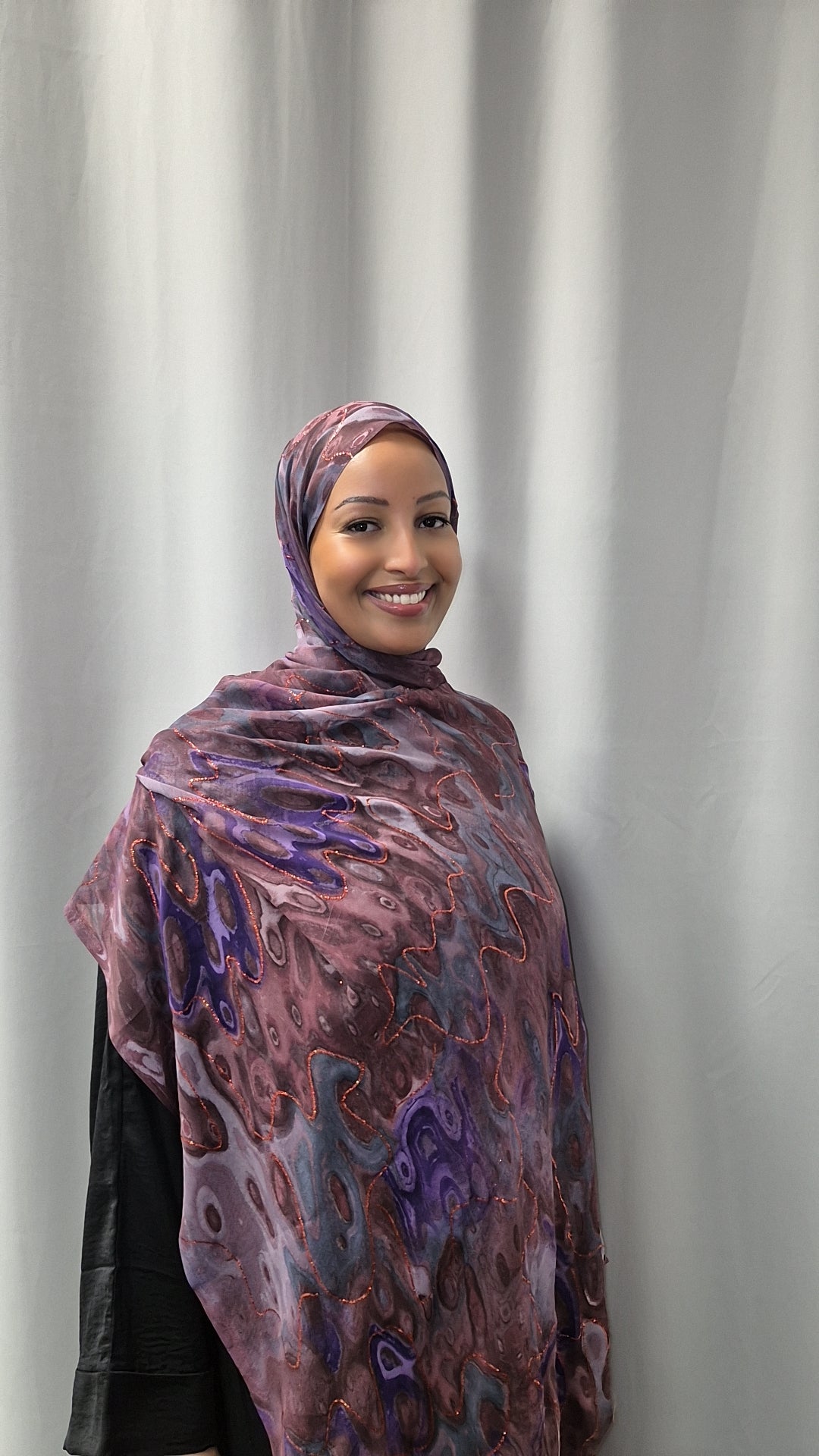 A woman in the Elegant Chiffon Hijab from Bihi Boutique smiles at the camera while standing before a plain light gray curtain.