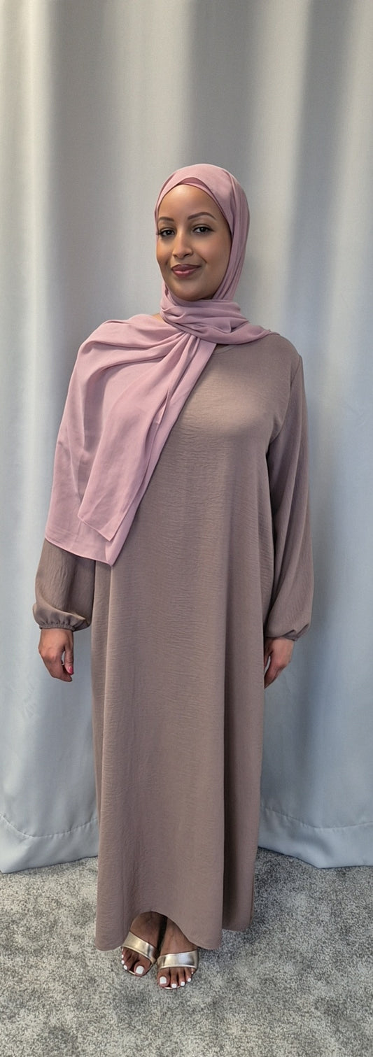 A woman stands in front of a light gray curtain, wearing the Shop Elegant Nude Slip Dress from Bihi Boutique. She pairs it with a matching pink hijab and silver open-toe sandals, smiling softly at the camera.