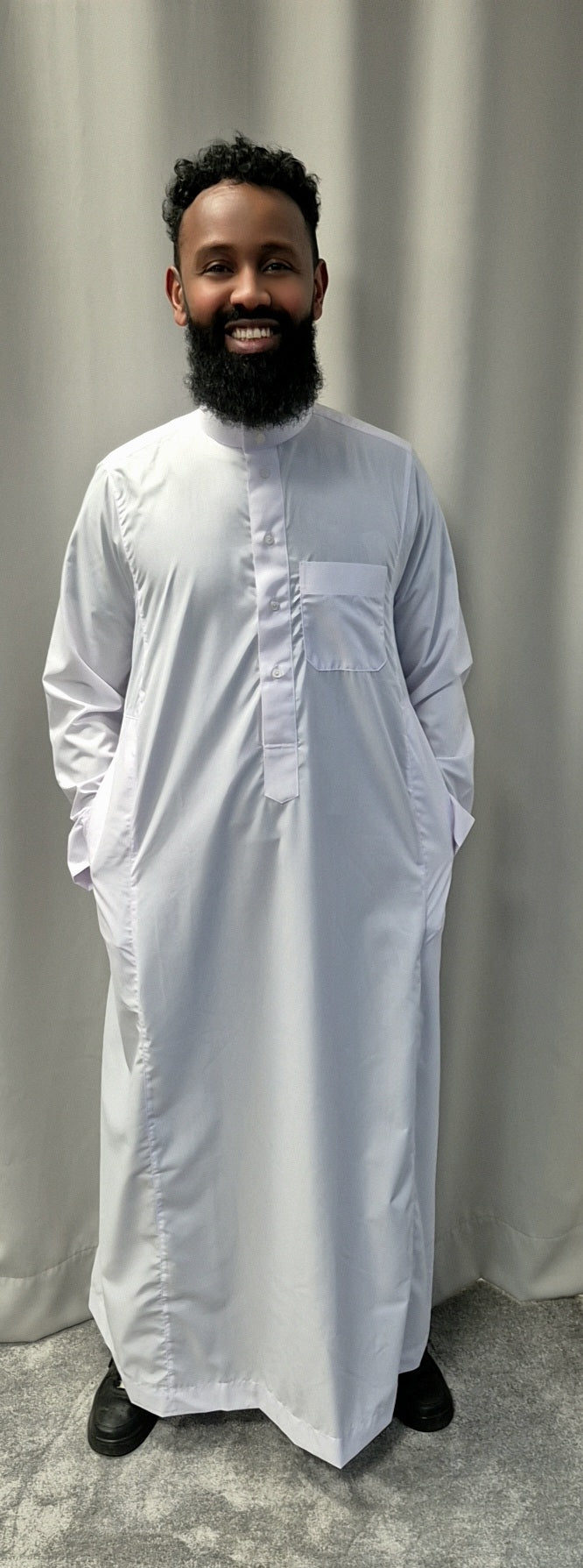 A bearded man smiles in front of a gray curtain, wearing the Shop Khamis Saudi White traditional attire from Bihi Boutique, paired with black shoes and his hands tucked in the garments pockets.