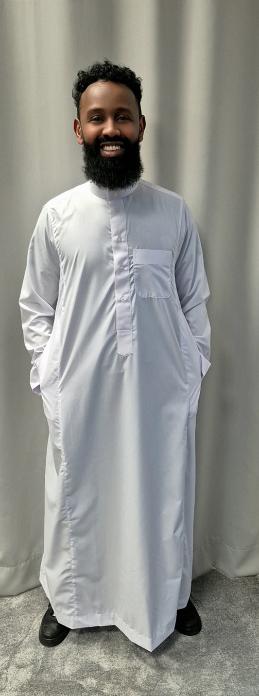 A bearded man smiles in front of a gray curtain, wearing the Shop Khamis Saudi White traditional attire from Bihi Boutique, paired with black shoes and his hands tucked in the garments pockets.
