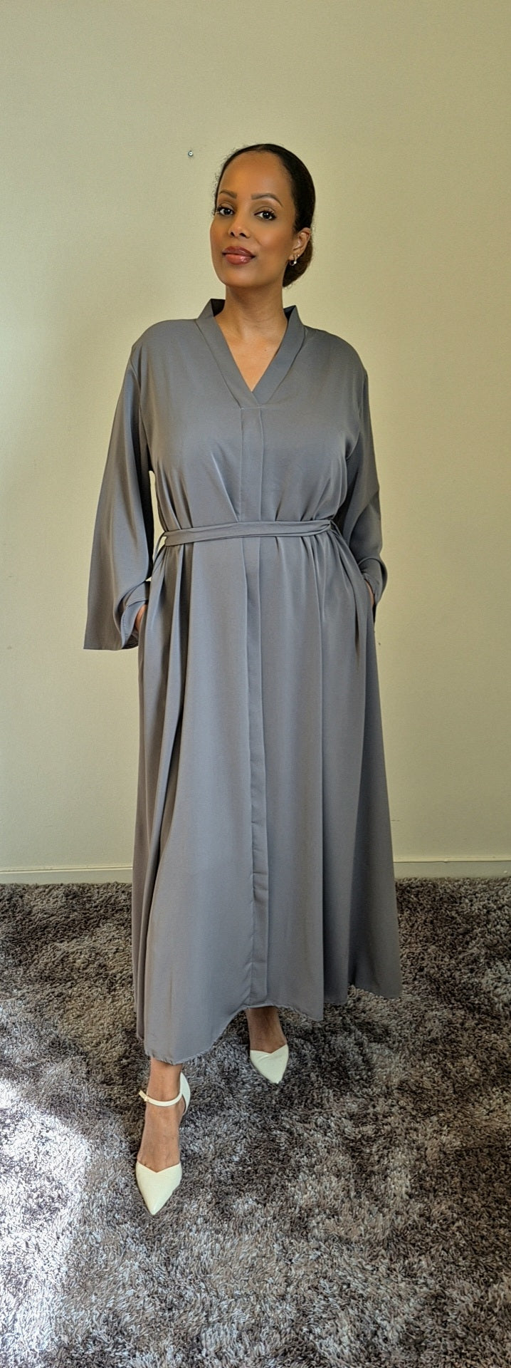 A woman stands on a carpeted floor wearing the Bihi Boutique Shop Layina Black Dress in L/XL, an elegant style with a belted waist, her hands in pockets, facing the camera against a plain light wall.