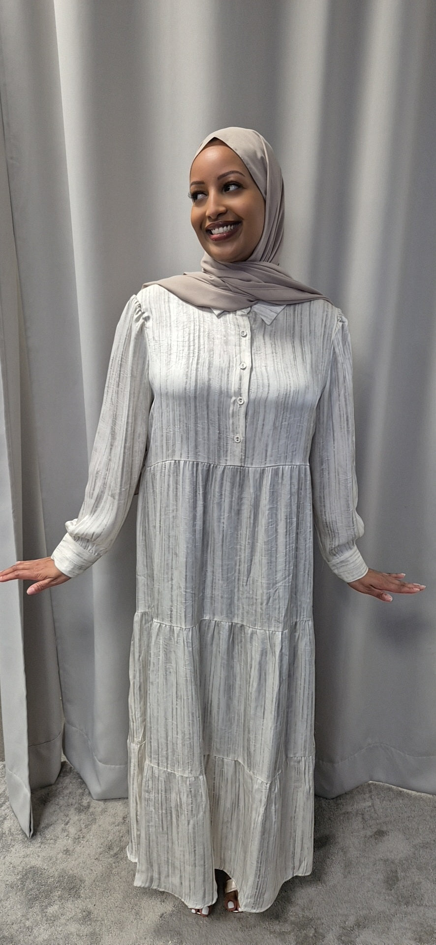 A smiling woman in the Celestial Ivory Satin Dress (Size S/M) by Bihi Boutique stands before a grey curtain, her arms relaxed at her sides, showcasing an elegant and chic look.