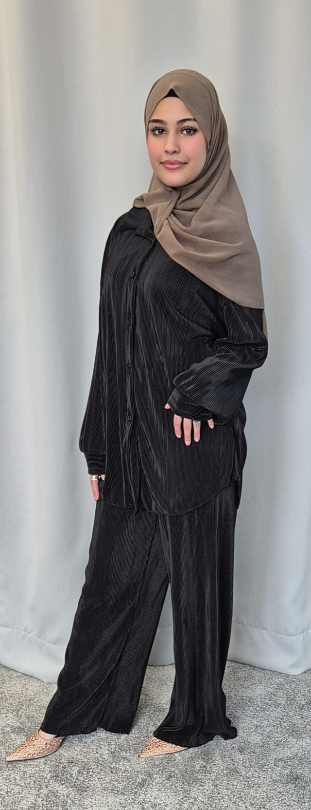 A woman in the Bihi Boutique Stylish Black Opal Co-ord Set, featuring a loose black pleated outfit and taupe hijab, smiles softly at the camera in front of a light gray curtain.