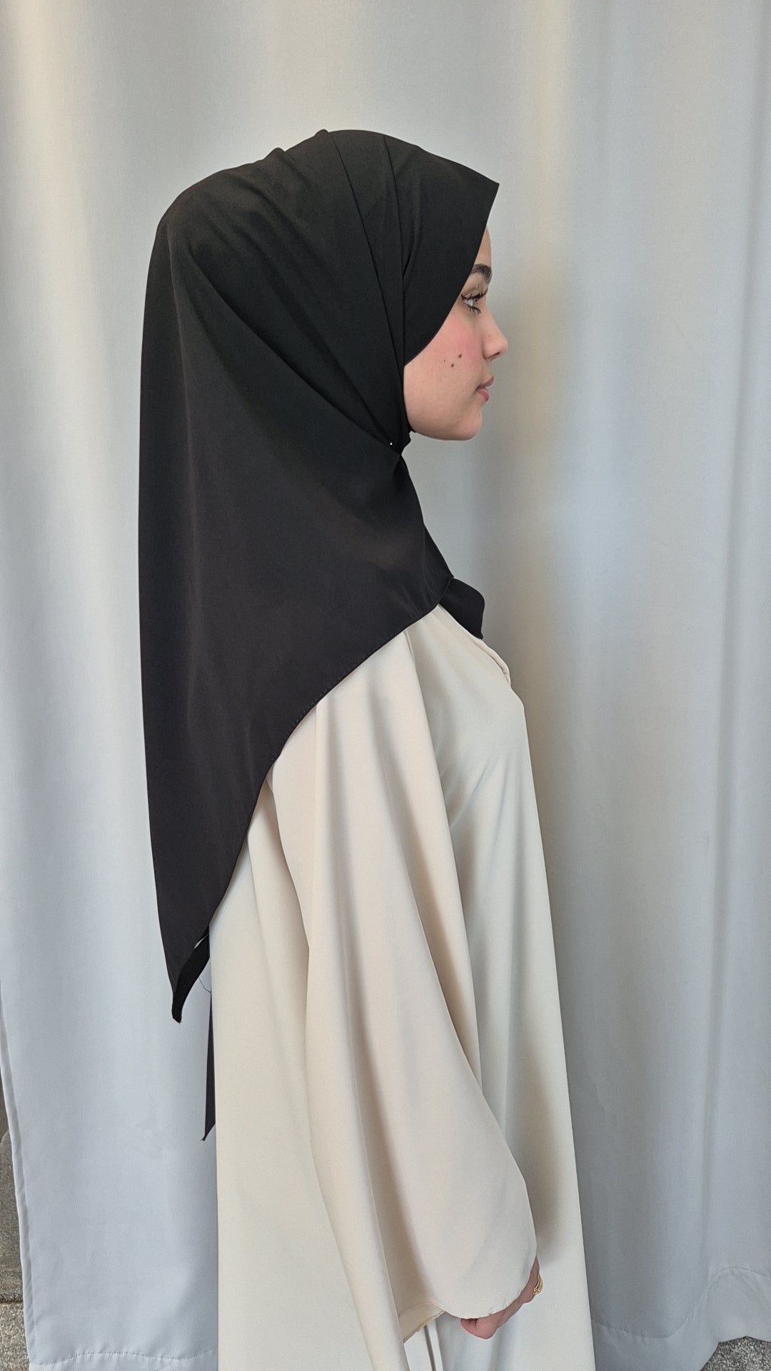 A woman in cream stands in profile against a light background, wearing the Shop Medina Seide Black 180x170 cm Blanket by Bihi Boutique as a draped hijab over her shoulders.