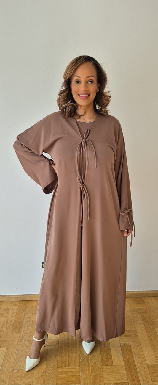A woman in the Stylish Ayan Brown Hijab Set by Bihi Boutique, made from luxurious Nidah and chiffon, stands smiling on a wooden floor against a light wall. She pairs the elegant outfit with white heels.