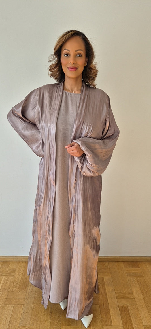 A woman stands indoors on a wooden floor, wearing the Elegant Organza Nude Slipdress & Chiffon Hijab in Size 58 by Bihi Boutique, smiling softly with one arm raised against a plain white wall.