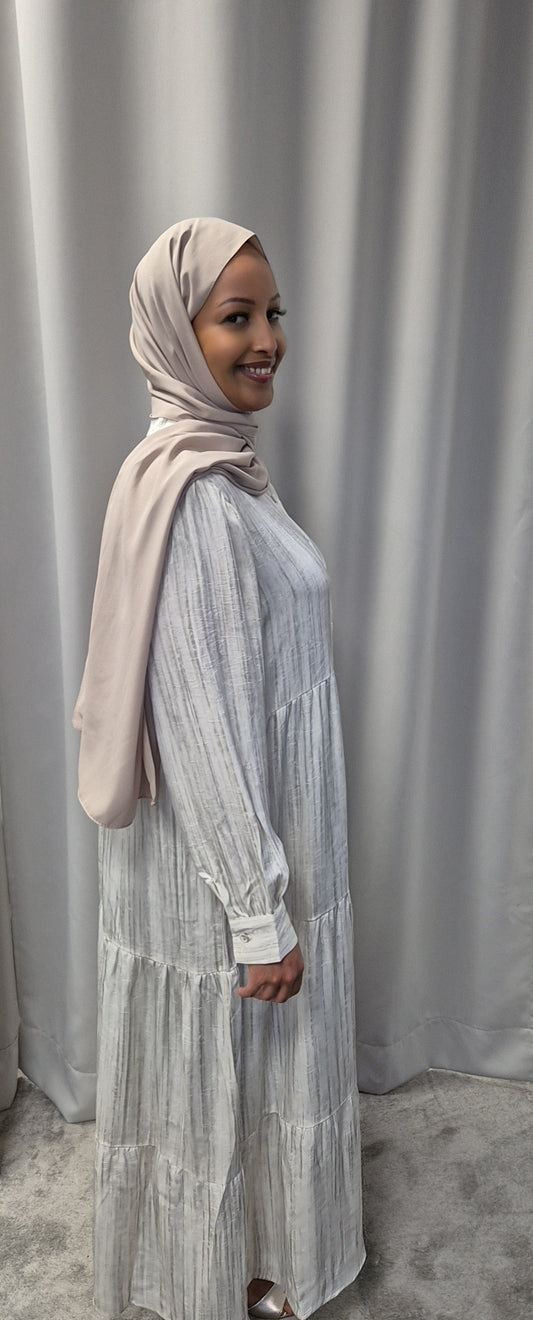 A woman in a light beige hijab wears the Bihi Boutique Celestial Ivory Satin Dress (Size S/M) for an elegant, chic look as she smiles in front of light gray curtains.