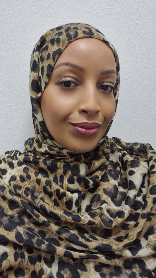 A woman models the Bihi Boutique Stylish Cheetah Hijab, smiling slightly at the camera while posing against a plain white wall. Elevate your fashion game today with this chic accessory!.