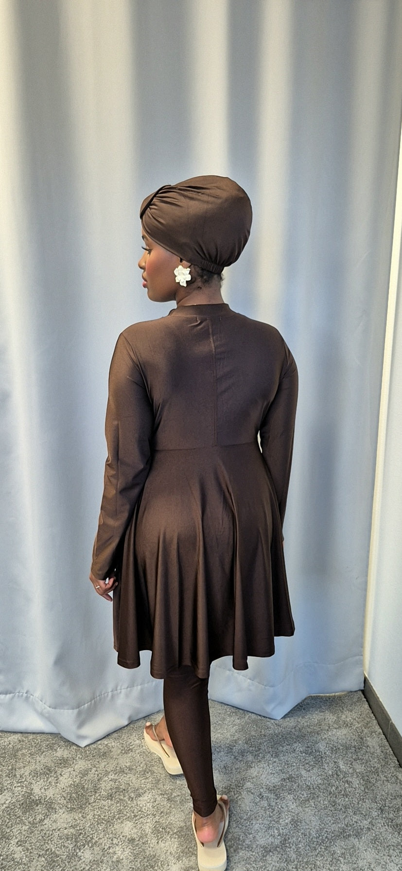 A person wearing the Shop Stylish Black Burkini by Bihi Boutique, featuring fitted sleeves and a flared skirt, stands with their back to a light gray curtain, accessorized with a matching headscarf and floral earrings.