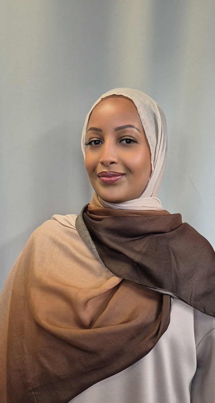 A woman models the Shop Stylish Ombre Hijab in beige and brown by Bihi Boutique, made from soft cotton and viscose, standing against a light gray background and smiling gently at the camera.