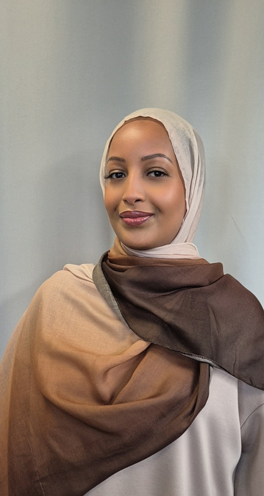 A woman models the Shop Stylish Ombre Hijab in beige and brown by Bihi Boutique, made from soft cotton and viscose, standing against a light gray background and smiling gently at the camera.
