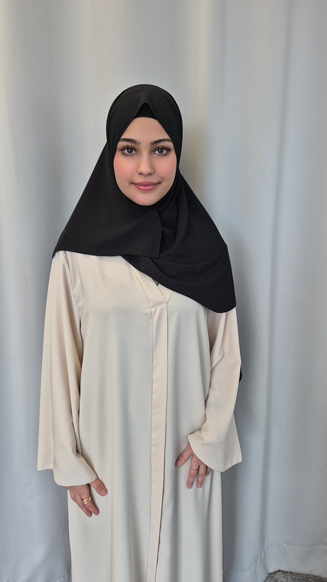 A young woman smiles gently at the camera, standing by a light curtain with her hands by her sides, wearing the Shop Medina Seide Black 180x170 cm blanket from Bihi Boutique.