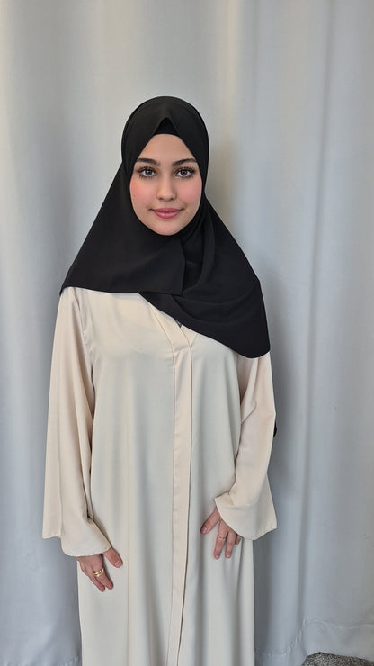 A young woman smiles gently at the camera, standing by a light curtain with her hands by her sides, wearing the Shop Medina Seide Black 180x170 cm blanket from Bihi Boutique.
