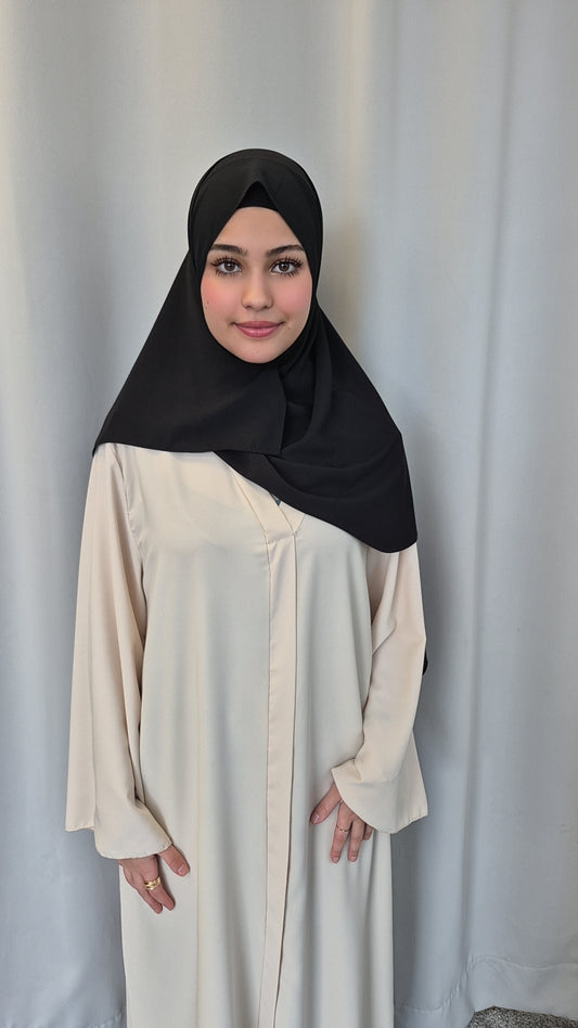 A young woman smiles gently at the camera, standing by a light curtain with her hands by her sides, wearing the Shop Medina Seide Black 180x170 cm blanket from Bihi Boutique.