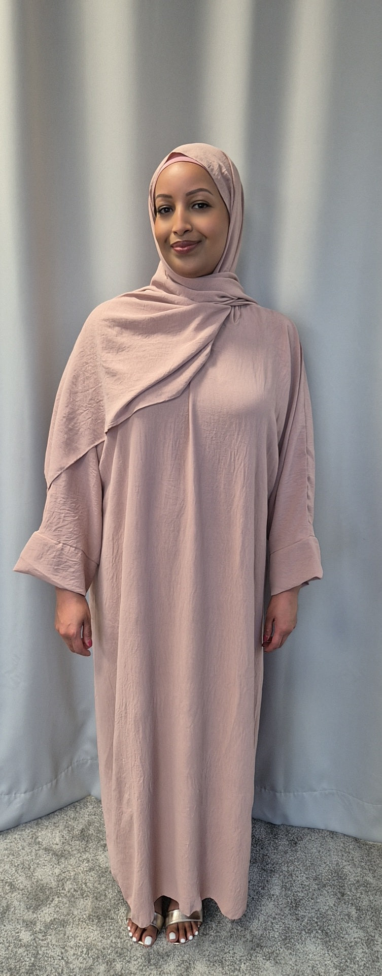 A woman wearing Bihi Boutiques Elegant Black One Size Prayer Clothing stands on a gray carpet before a light gray curtain, smiling softly at the camera.