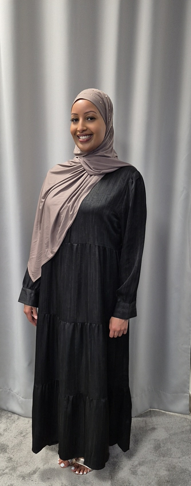 A woman wears the Bihi Boutique Shop Celeste Noir & Ivory Satin Dress (sizes S/M & L/XL) and a taupe hijab, smiling in front of light gray curtains.