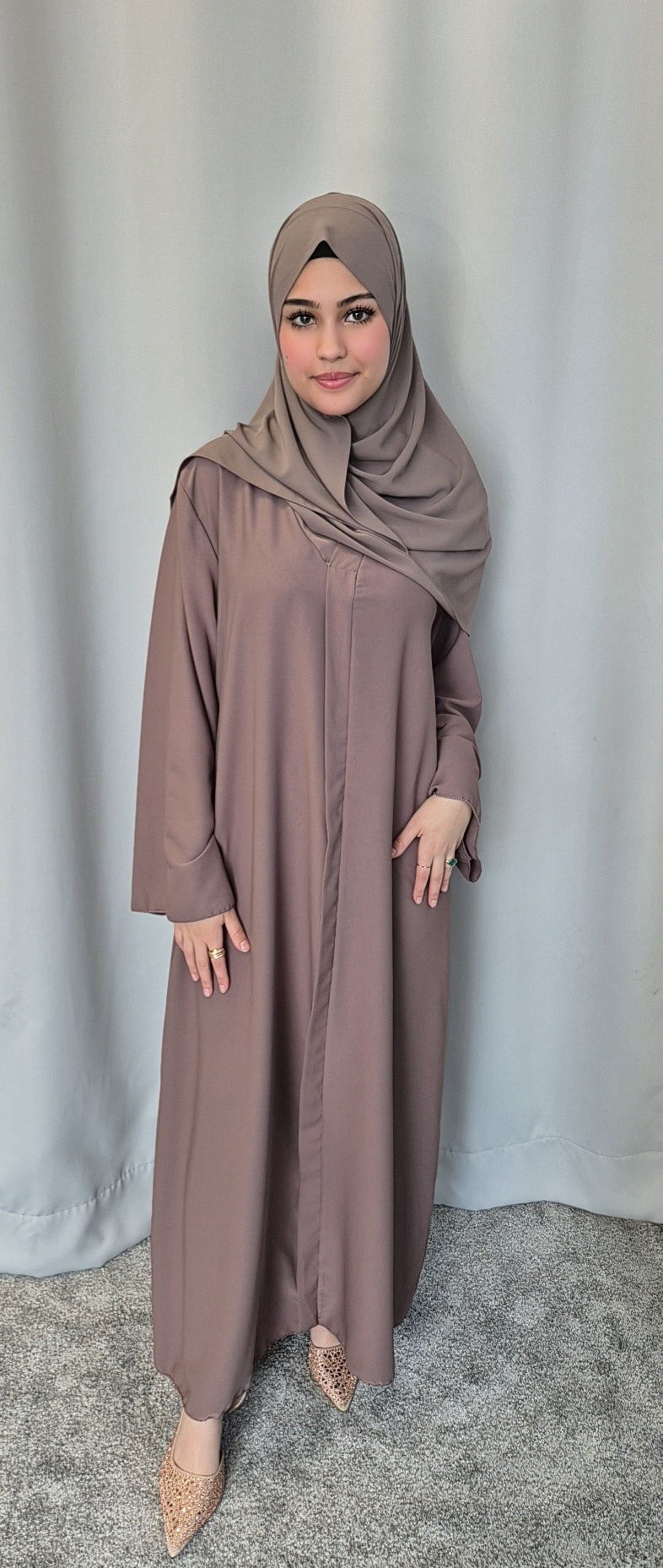 A woman wears the Islene Dress in taupe by Bihi Boutique with a matching hijab and light shoes, standing on a gray carpet before pale curtains, smiling softly. The one-size dress fits 160-165 cm and comes in 3 colors.