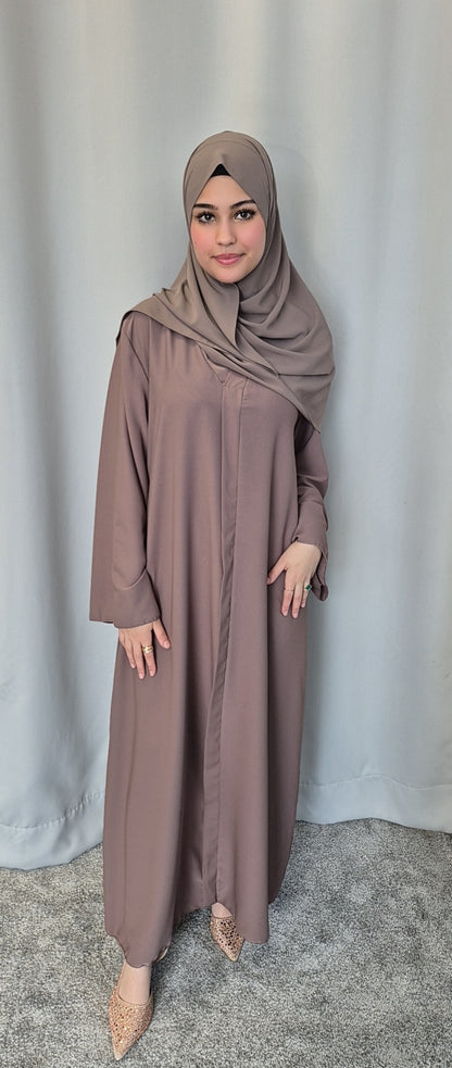 A woman wears the Islene Dress in taupe by Bihi Boutique with a matching hijab and light shoes, standing on a gray carpet before pale curtains, smiling softly. The one-size dress fits 160-165 cm and comes in 3 colors.