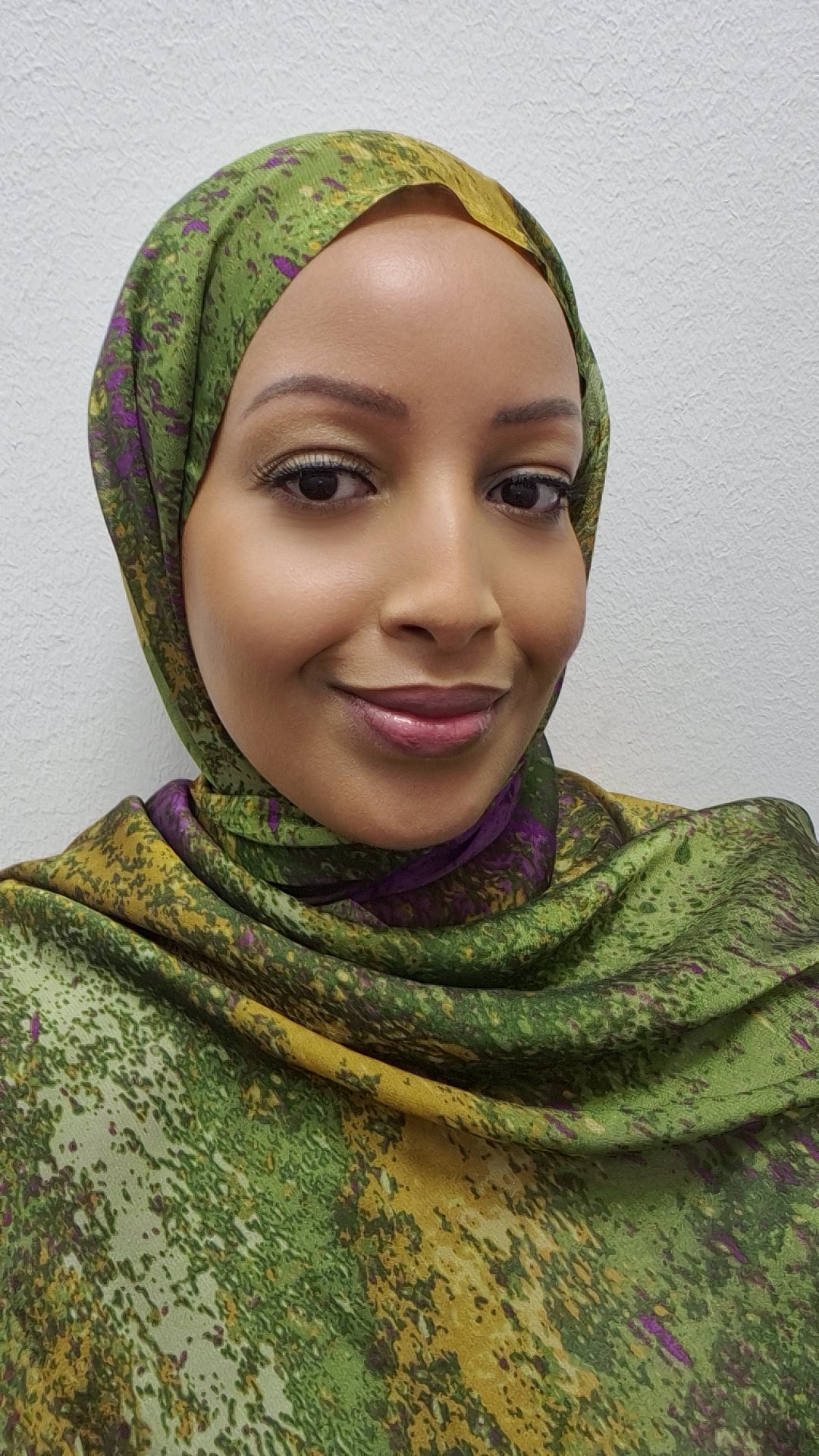 A woman models the Elegant Chiffon Hijab by Bihi Boutique, featuring a green, yellow, and purple pattern. She smiles gently at the camera, standing against a plain white background.