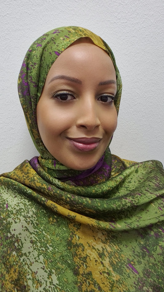 A woman models the Elegant Chiffon Hijab by Bihi Boutique, featuring a green, yellow, and purple pattern. She smiles gently at the camera, standing against a plain white background.