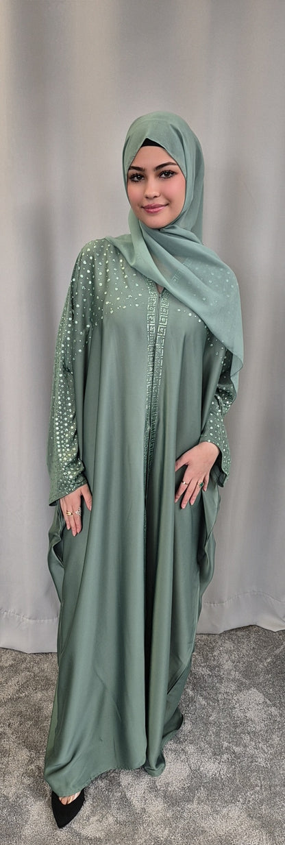 A woman smiles at the camera, wearing the Stylish Nourah Dress by Bihi Boutique in sage green with pearl-embellished sleeves and chest, plus a matching hijab. She stands on a gray carpet before a light gray curtain. Perfect Fit Size 52 & 54.
