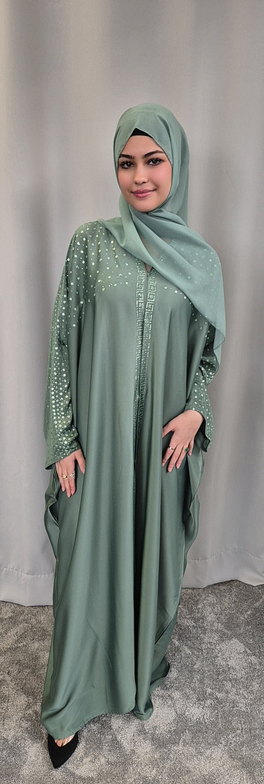 A woman smiles at the camera, wearing the Stylish Nourah Dress by Bihi Boutique in sage green with pearl-embellished sleeves and chest, plus a matching hijab. She stands on a gray carpet before a light gray curtain. Perfect Fit Size 52 & 54.