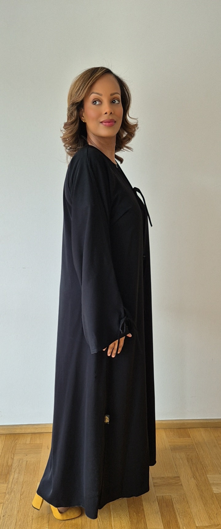A woman with shoulder-length hair stands sideways indoors on a wooden floor, wearing the Bihi Boutique Ayan Black Nidah Dress with Chiffon Hijab in Size 60. She faces left, looking at the camera with a slight smile.