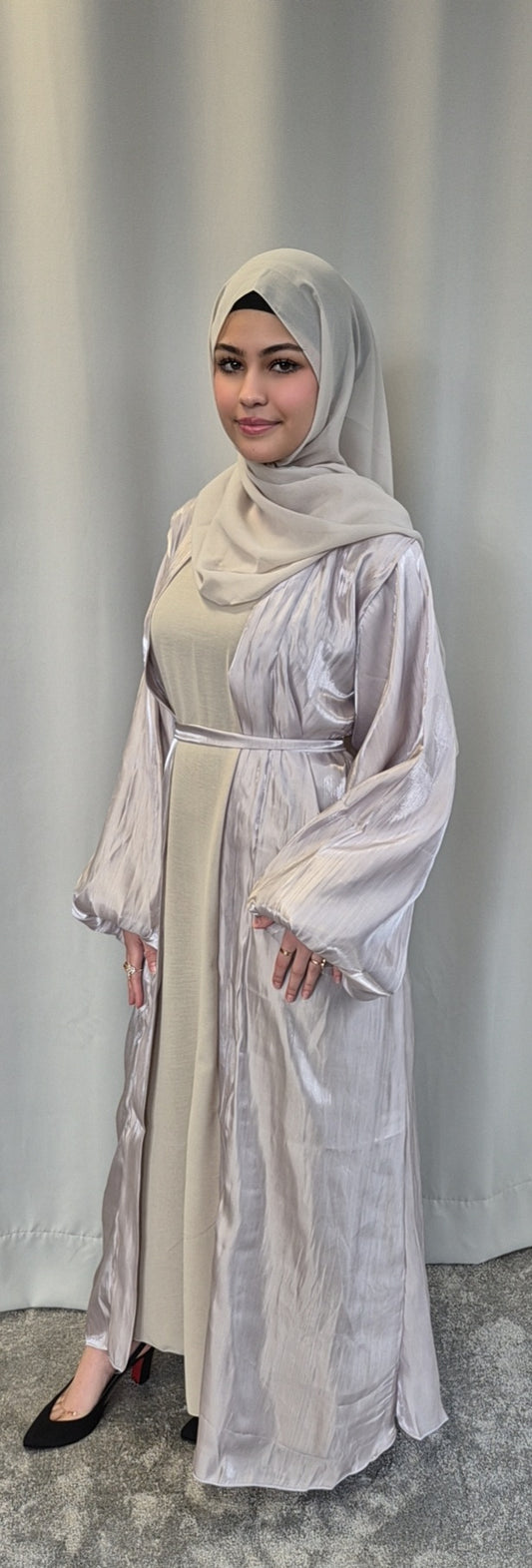A woman smiles in front of a light gray curtain, wearing the Bihi Boutique Stylish Organza Chrome Hijab Set—a chic, lightweight beige hijab with a matching long dress and sheer outer layer—paired with black heels.