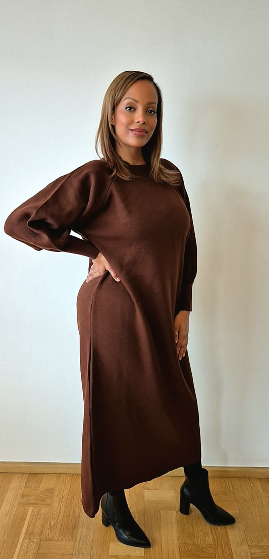 A woman poses indoors against a white wall, wearing the Bihi Boutique Chic One Size Brown Sweater Dress—made from a cozy wool blend—with long sleeves and black ankle boots. She smiles slightly and stands on a wooden floor.