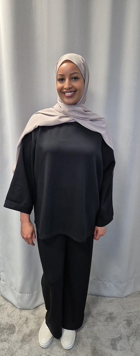 A woman smiles in front of a light grey curtain, wearing Bihi Boutiques Calma Luxe Nue & Noir one size fits all hijab, styled with a loose black top, black pants, and white shoes.