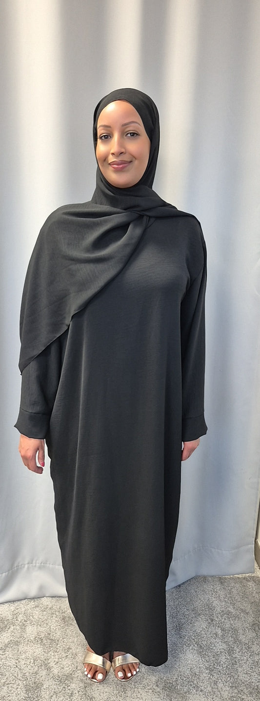 A woman smiles at the camera in front of a light gray curtain, wearing Bihi Boutique’s Elegant Black One Size Prayer Clothing for All Sizes and open-toed sandals.